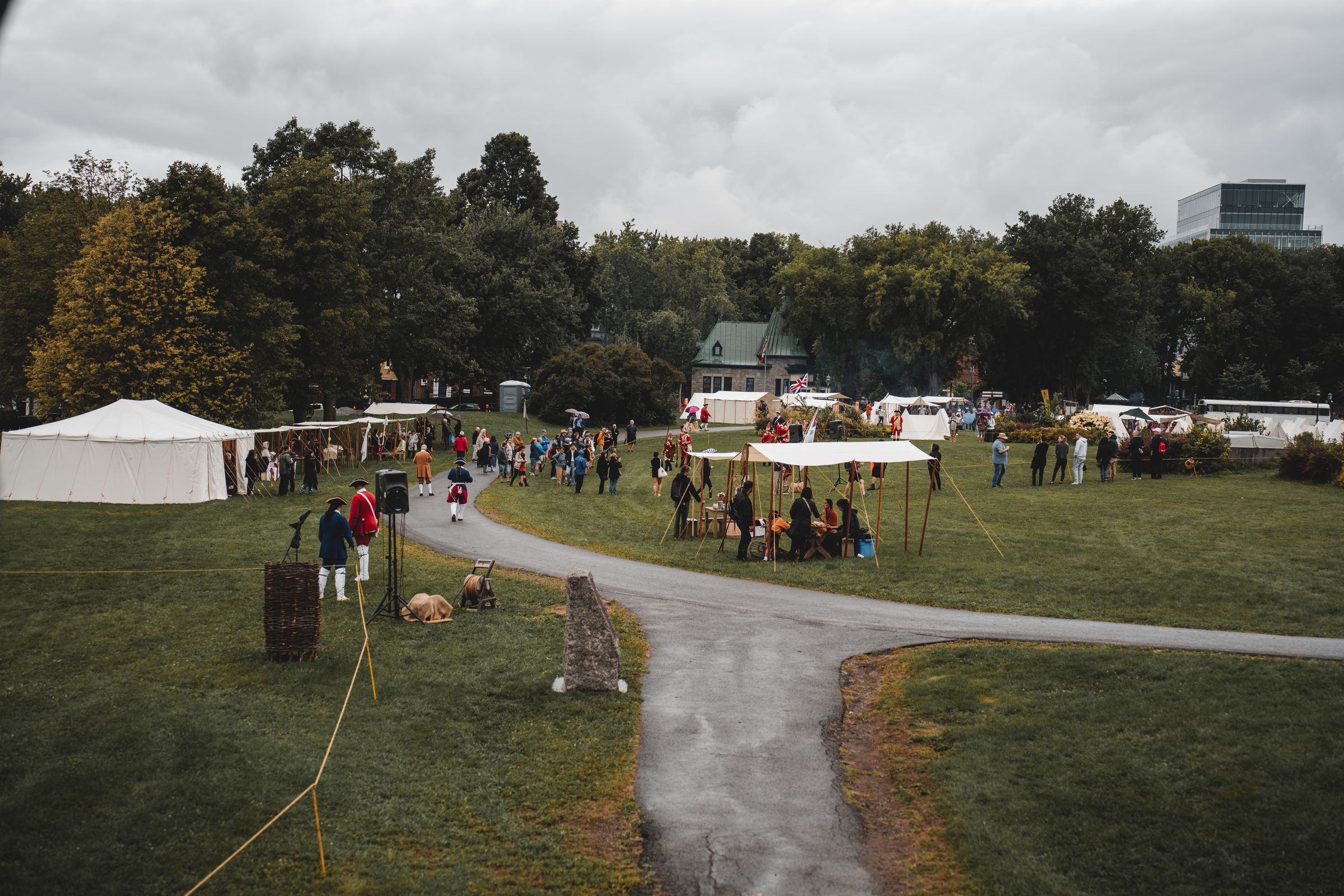 A community day laid out across an open park field: rows of white canvas tents, a path curving through the grass, and people gathered in the open under a grey sky.